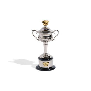 A miniature replica Women's AO trophy with a silver cup, two handles, and a gold-colored tennis rackets on top, presented on a black base.