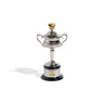 A miniature replica Women's AO trophy with a silver cup, two handles, and a gold-colored tennis rackets on top, presented on a black base.
