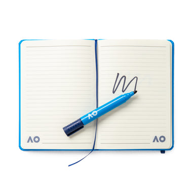 An open lined notebook with a blue marker pen placed across the pages. The pen has made a black squiggly mark on the right-hand page. Both pages feature a small "AO" logo in the bottom corners, and the notebook has a blue cover and ribbon bookmark.