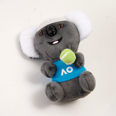 Side view of a plush toy koala with a white headband and a blue polo shirt with the AO logo on it, holding a tennis ball.